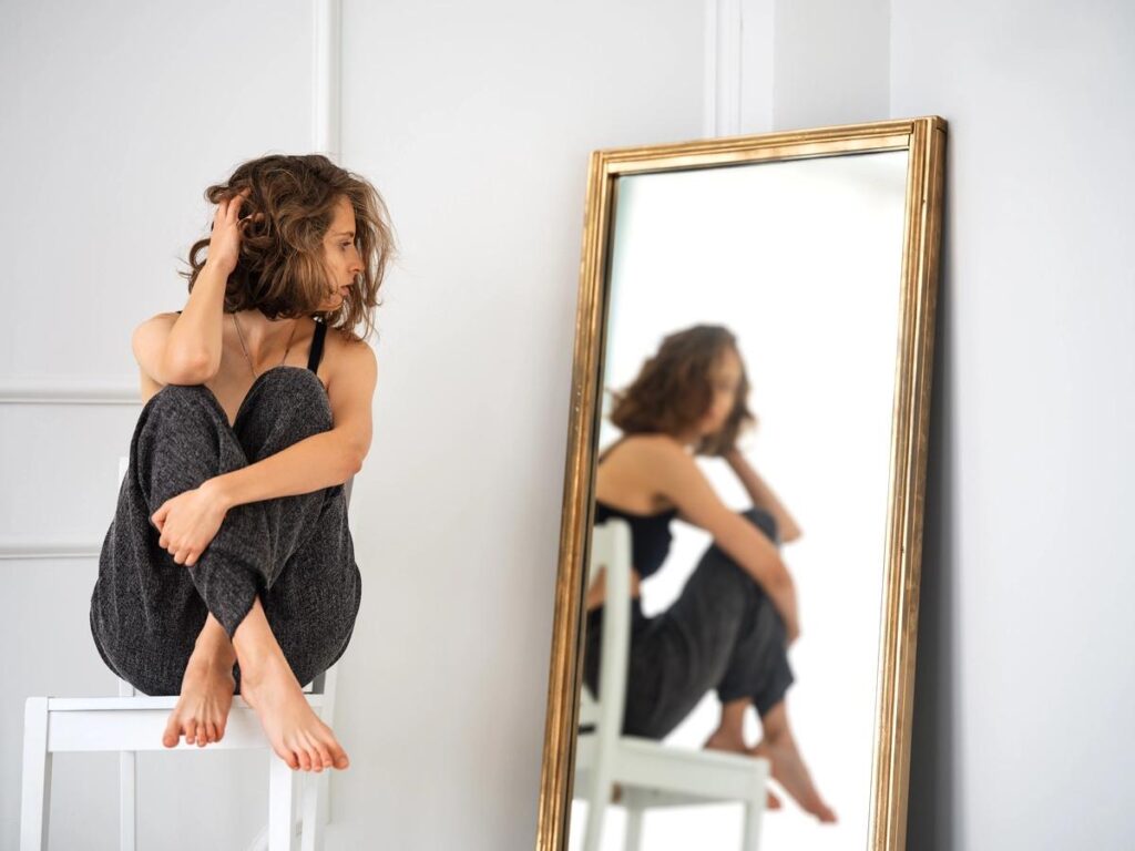 Mulher sentada diante do espelho, refletindo sobre expectativas e autocobrança.
